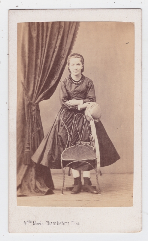 Jeune fille tenant son chapeau, posant debout derrière une chaise 