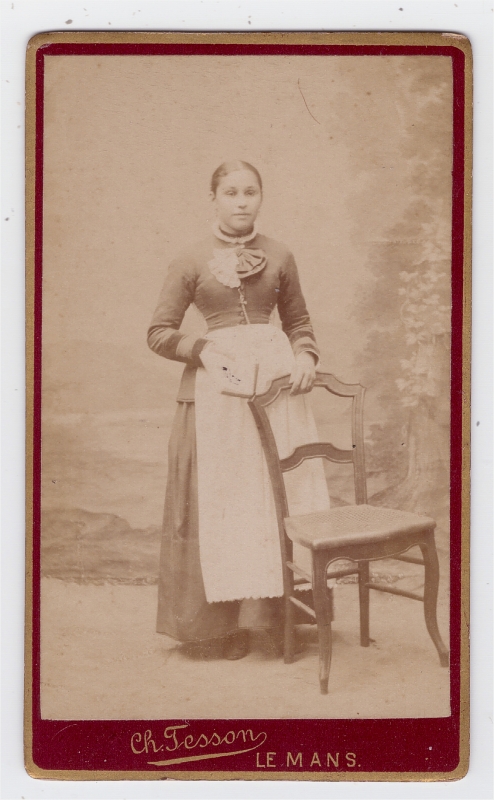 Jeune femme en tablier (domestique ?) posant debout derrière une chaise